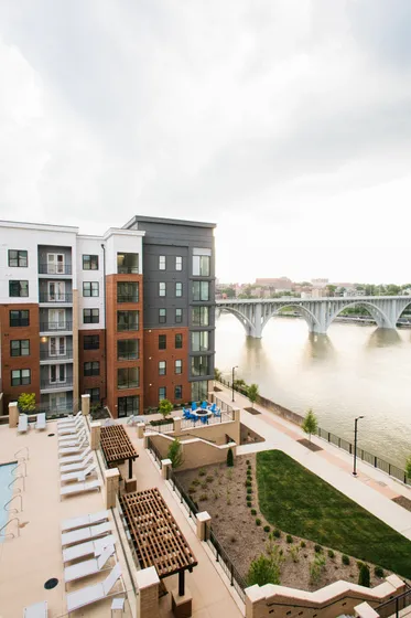 Modern riverside apartment complex with stunning architecture, featuring a scenic view of the bridge and river, and inviting outdoor amenities including lounge chairs and vibrant patio seating.