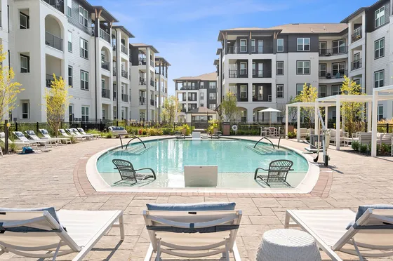 Luxurious apartment complex showcasing a stunning pool area with elegant lounge chairs and lush landscaping under a bright blue sky.