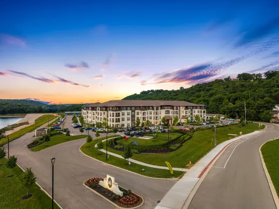 Aerial view of a luxurious apartment complex nestled in a picturesque landscape with vibrant greenery and a serene lake, under a stunning sunset sky.