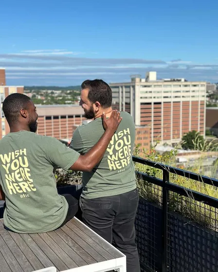 Men enjoying a sunny rooftop with stunning cityscape views, wearing matching 'Wish You Were Here' shirts.