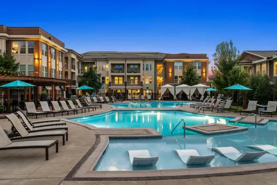 Elegant apartment complex with a sparkling pool, lounge chairs, and cabanas under a vibrant evening sky.
