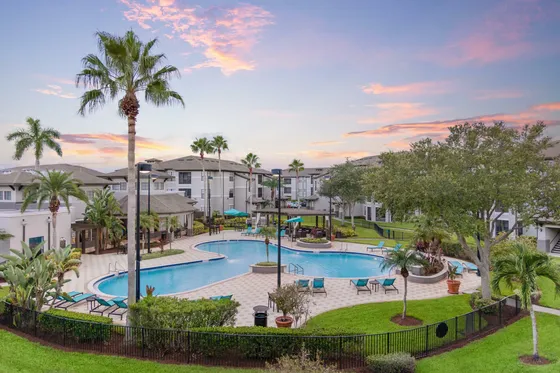 Inviting community pool area surrounded by palm trees and modern apartments, with a backdrop of a stunning sunset sky.