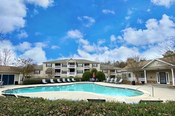 Charming apartment complex with a serene swimming pool, surrounded by lush greenery and inviting lounge chairs under a bright blue sky.