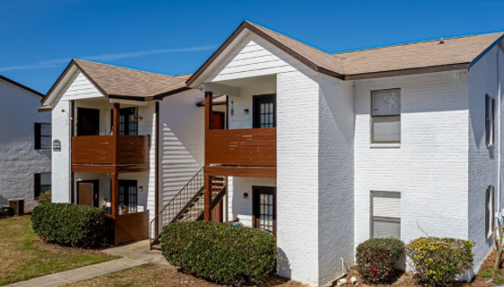 Charming white brick apartment complex with wooden balconies, surrounded by well-maintained bushes and a clear blue sky.
