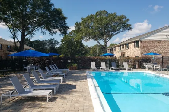 Inviting outdoor pool area with modern lounge chairs and vibrant blue umbrellas, surrounded by lush greenery and charming brick buildings under a clear sky.