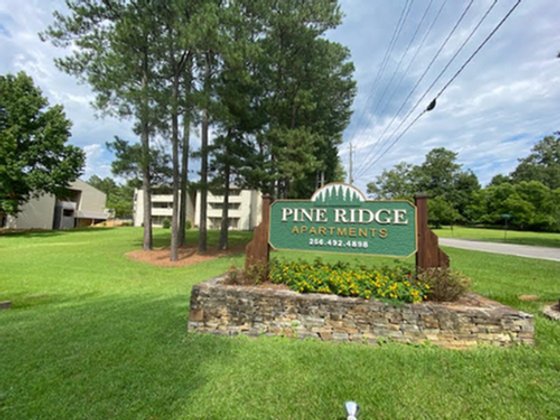 Inviting entrance to Pine Ridge Apartments surrounded by lush greenery and tall trees, offering a serene living environment.