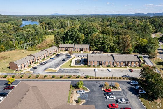 Aerial view of a charming apartment complex surrounded by lush greenery and scenic landscapes, with ample parking and easy access to serene walking paths by the lake.