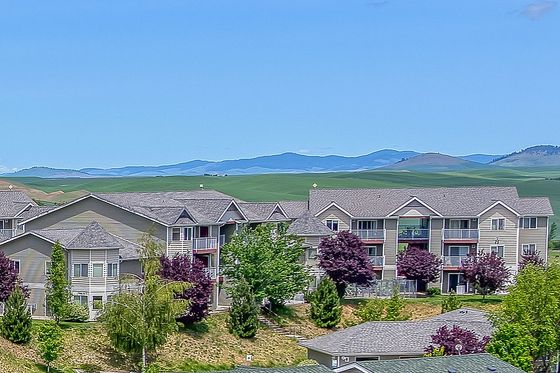 Charming residential complex with multiple apartment buildings, green lawns, and ample parking set against a backdrop of rolling hills and a clear blue sky.