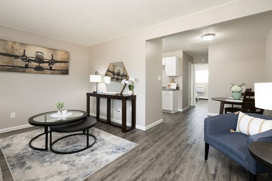 Chic living room with modern decor including a sleek glass coffee table, elegant artwork, and cozy seating leading into a bright kitchen area.
