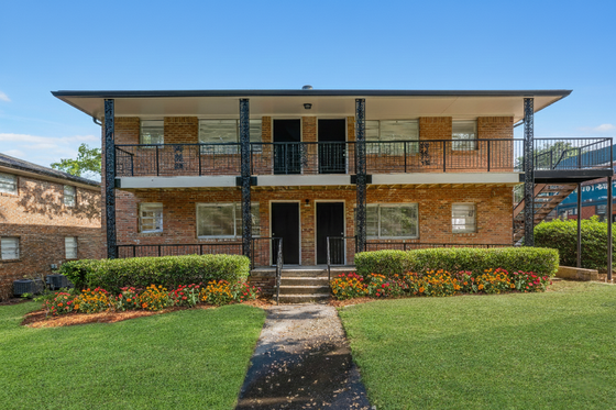 Charming two-story brick apartment building surrounded by lush greenery and vibrant flowerbeds, offering a warm and inviting residential ambiance.