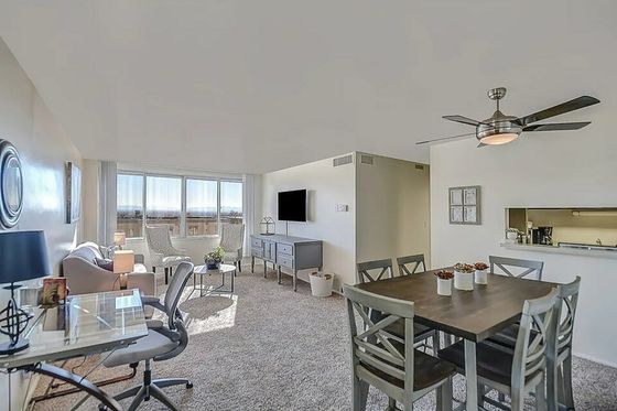 Contemporary open-plan living area with dining set, plush seating, and a ceiling fan. Sunlight streams through large windows.