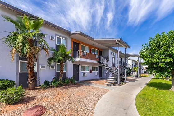 Modern two-story apartment building with vibrant palm trees, lush green surroundings, and a clear blue sky.