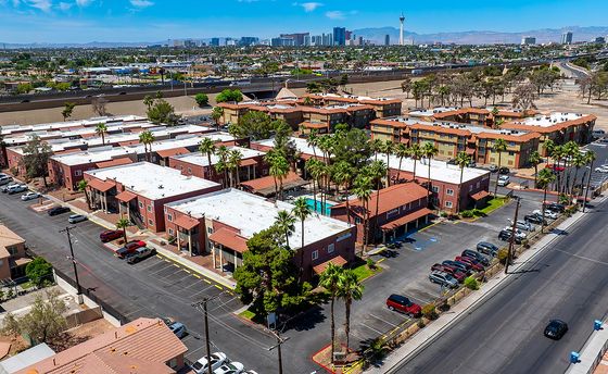 Vibrant aerial view of charming apartment complex with lush palm trees, nestled near bustling cityscape under clear blue skies.