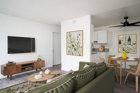 virtually staged living room with faux hardwood flooring and a green sofa with two coffee tables in front an entertainment console with a tv on the wall and a kitchen with a round dining table at boulder gardens