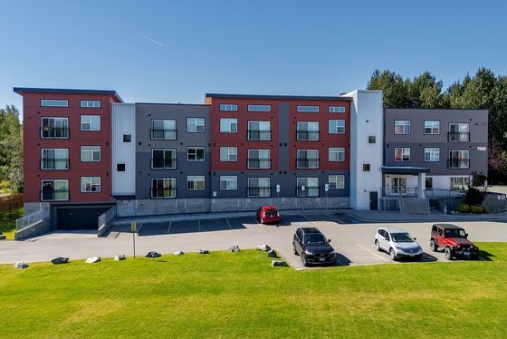 Modern apartment building with colorful facade and large windows, surrounded by lush greenery and a spacious parking area.