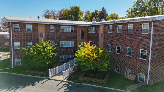 Charming brick apartment building with lush green trees and a welcoming pathway, perfect for a cozy living experience.
