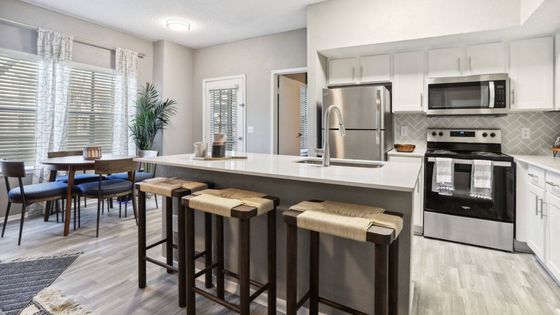 Modern kitchen with stainless steel appliances, white quartz island, wooden bar stools, vinyl flooring, and a large houseplant