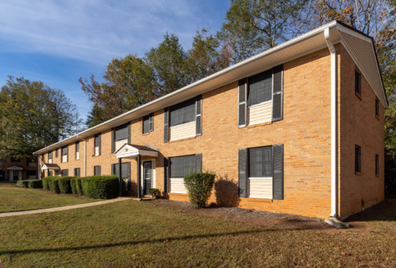 Charming brick apartment building with classic black shutters, nestled in a lush green setting and bathed in warm sunlight.