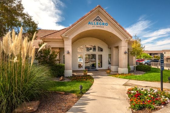 Inviting entrance of Allegro at Tanoan, surrounded by lush greenery and vibrant flowers, with a charming architectural design under a clear blue sky.