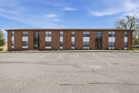 Charming two-story red brick apartment building with modern design accents and spacious front parking area under a clear blue sky.