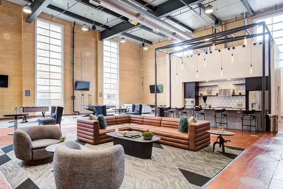 Modern loft-style communal area with high ceilings, industrial design, and versatile seating. Features include sleek kitchen island, plush sofas, cozy chairs, and ample natural light from tall windows.