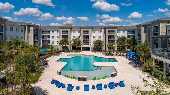 Elegant apartment complex featuring a stunning outdoor pool surrounded by lush palm trees and stylish lounging areas, set against a backdrop of modern architecture and a clear blue sky.