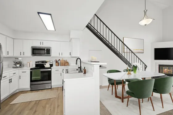 Modern kitchen and dining area showcasing sleek white cabinetry, stainless steel appliances, and a chic dining table with elegant green velvet chairs, next to a staircase and a cozy fireplace.