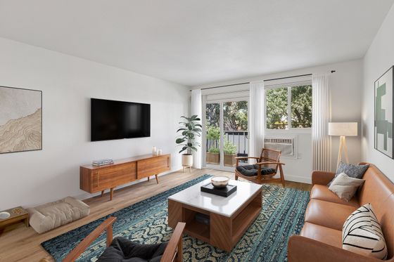 Modern living room with sleek decor, featuring a stylish tan leather sofa, mid-century wooden furniture, and a vibrant patterned rug. Natural light floods through large windows, highlighting greenery on the balcony.