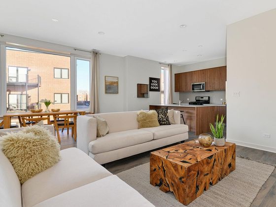 Bright and modern open-concept living room with cozy seating, stylish wooden coffee table, and sleek kitchen with stainless steel appliances. Large windows illuminate the space with natural light.
