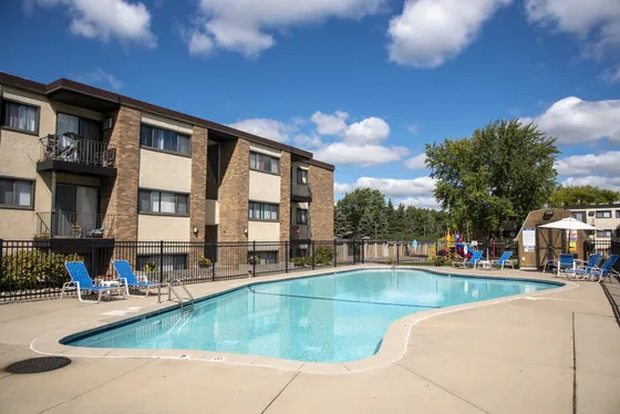 Inviting apartment building with a sparkling outdoor pool, surrounded by lush greenery and vibrant blue lounge chairs, under a clear blue sky.