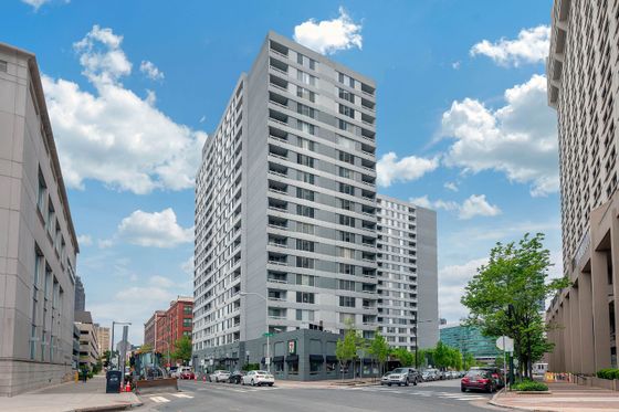 Modern high-rise apartment building with sleek design, situated on a bustling city street with lush greenery and clear blue skies.