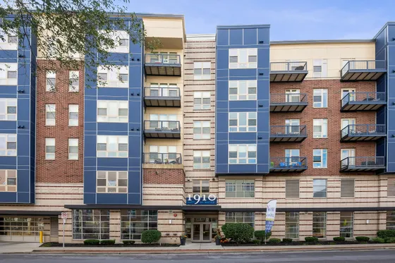 Modern apartment complex with striking blue and brick exterior, featuring private balconies and lush greenery at the entrance.