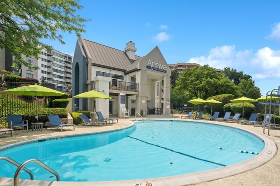 Inviting apartment community pool area featuring modern architecture and vibrant green umbrellas, set against a backdrop of lush trees and residential buildings.