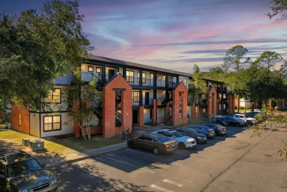 Charming apartment complex at sunset with modern brick and wood design, lush trees, and ample parking.