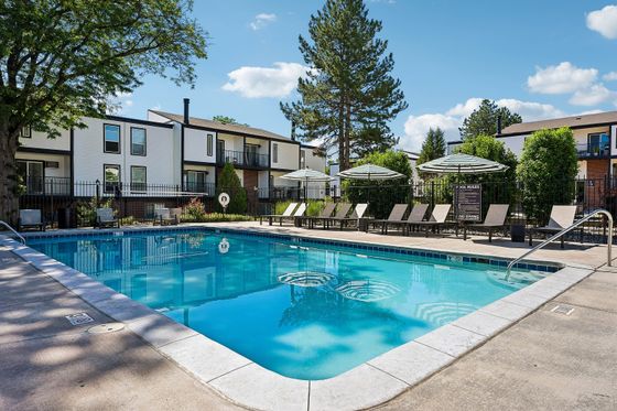 Inviting apartment pool area with sun loungers, umbrellas, and lush landscaping, set against a backdrop of modern two-story buildings.