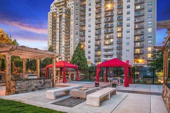Chic urban apartment complex with illuminated modern high-rise, vibrant outdoor seating areas, red canopies, and a stylish stone pergola against a stunning sunset backdrop.