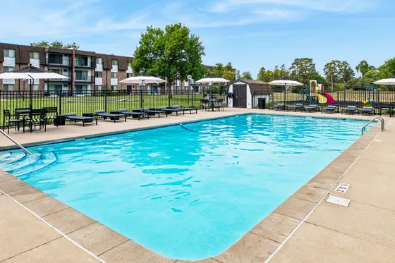 Inviting outdoor pool area with sun loungers and umbrellas, perfect for relaxation. Scenic backdrop of modern apartment buildings and a colorful playground, ideal for family living.