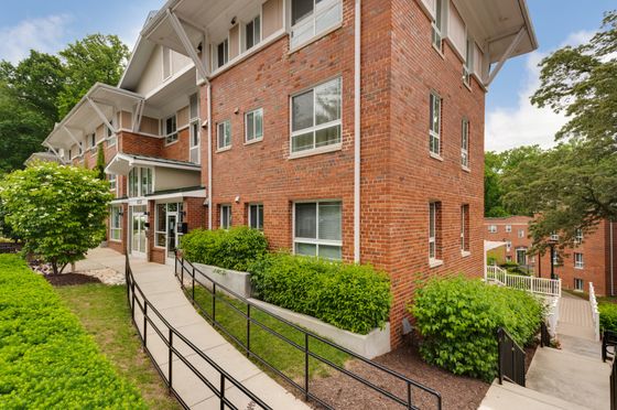 Charming brick apartment building surrounded by lush greenery, featuring large windows and a welcoming entrance path.