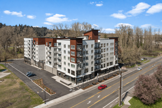 Modern apartment complex with stylish architecture, ample parking, and convenient road access, set against a picturesque wooded backdrop.