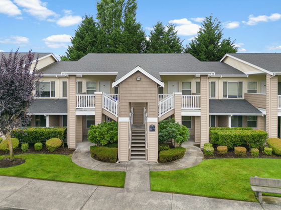 Charming two-story apartment building with neatly trimmed landscaping and inviting front balconies, set against a backdrop of lush trees and a clear blue sky.