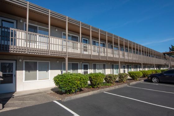 Charming two-story apartment building with modern balconies, lush greenery, and ample parking space under a bright blue sky.