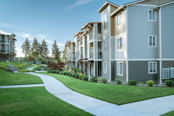 Charming modern apartment complex with lush green lawns and a scenic walking path, surrounded by trees under a clear blue sky.