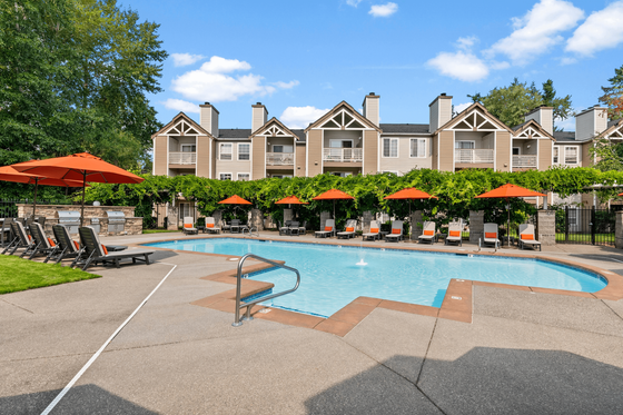 Inviting apartment complex boasting a sparkling swimming pool, comfortable lounge chairs under vibrant orange umbrellas, and charming architecture enveloped in lush greenery.