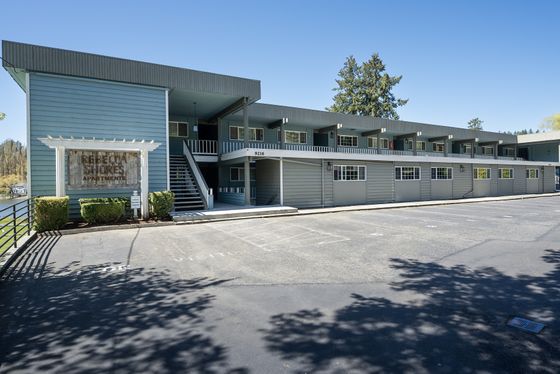Charming two-story apartment complex with stylish blue siding, nestled in a serene setting with lush greenery and ample parking space.