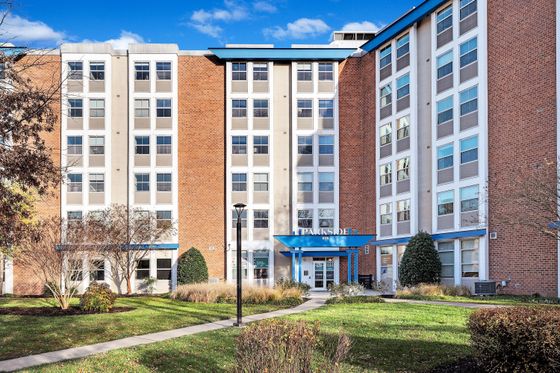 Modern apartment complex exterior featuring a charming red brick facade, abundant windows, and a welcoming entrance surrounded by lush green landscaping.