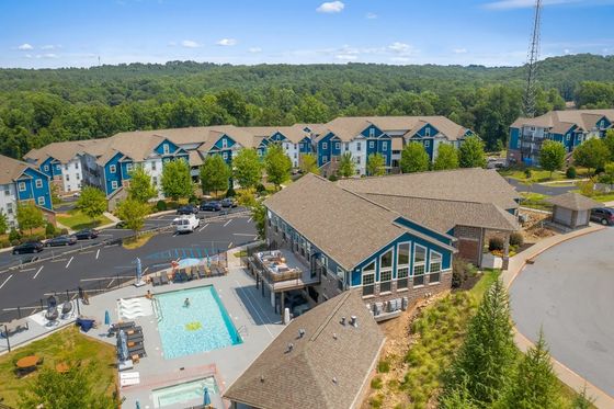 Aerial view of a serene apartment complex with modern blue-roofed buildings, surrounded by lush greenery. Features a sparkling swimming pool and spacious deck area, perfect for relaxation and outdoor enjoyment.