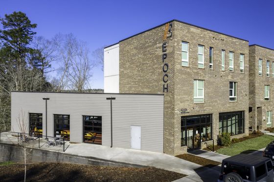 Modern three-story brick building with large windows, exterior signage, and inviting outdoor seating area under a clear blue sky.