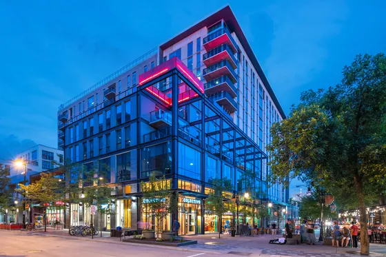 Vibrant modern urban building with glass facade and distinct red accents illuminated against the evening sky, surrounded by lively street life and lush greenery.