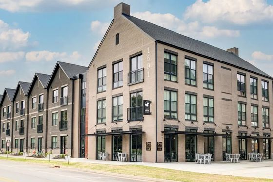 Modern three-story apartment building with stylish brick facade and large windows, showcasing urban living with outdoor seating on a sunny day.