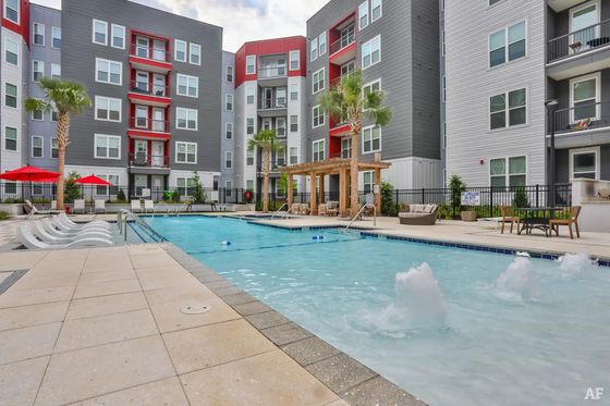 Modern apartment complex with a luxurious outdoor pool area, featuring sleek grey and red building design, palm trees, and inviting lounge spaces.